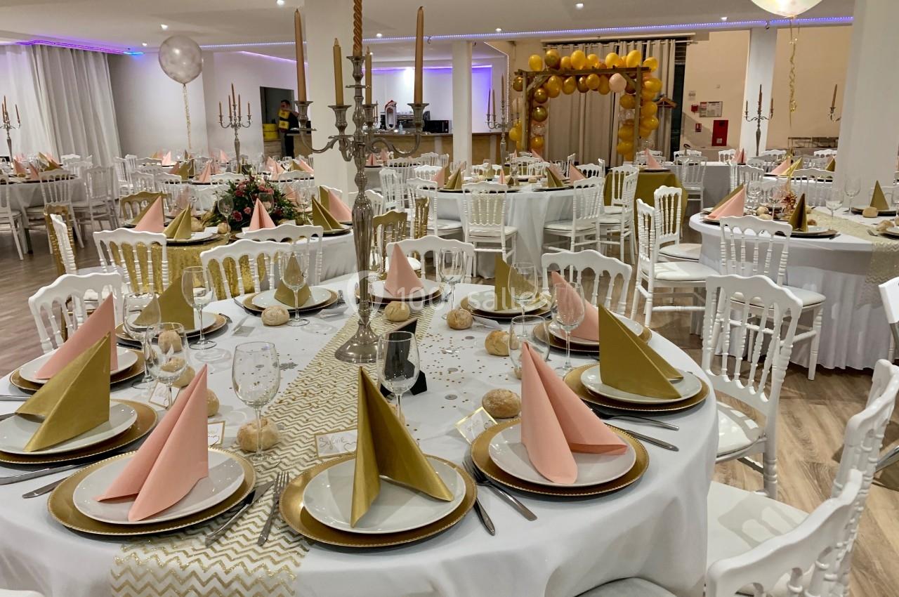 Salle de réception décorée avec des tables rondes dressées, nappes blanches, serviettes pliées et ballons dorés en arrière…