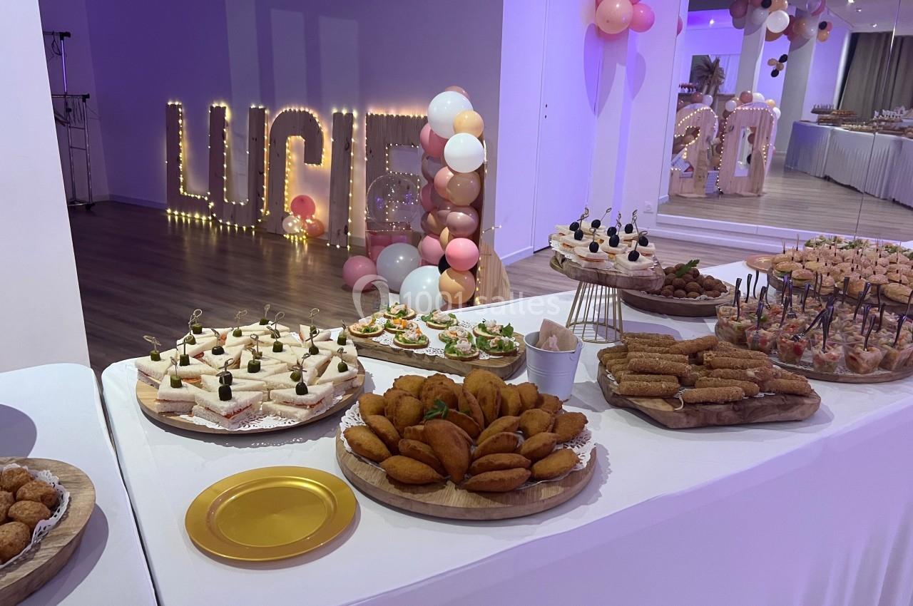 Buffet de réception avec divers amuse-bouches et décorations festives, dont des ballons et lettres lumineuses.