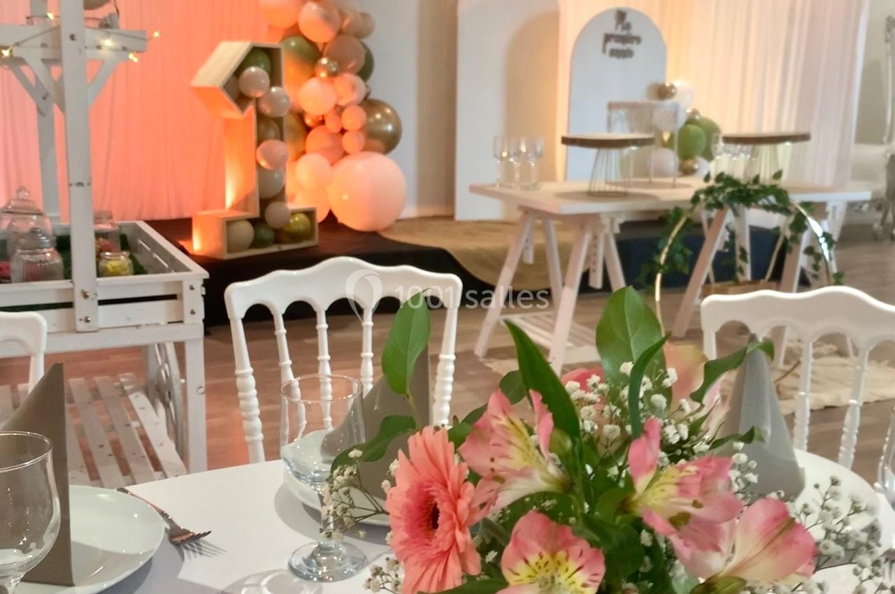 Salle décorée pour un événement, avec ballons, fleurs sur table et mobilier blanc dans une ambiance lumineuse.