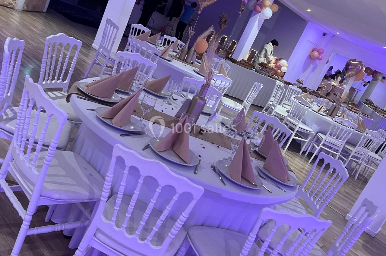 Salle décorée pour un événement avec tables rondes, chaises blanches, serviettes pliées et ballons roses en arrière-plan.