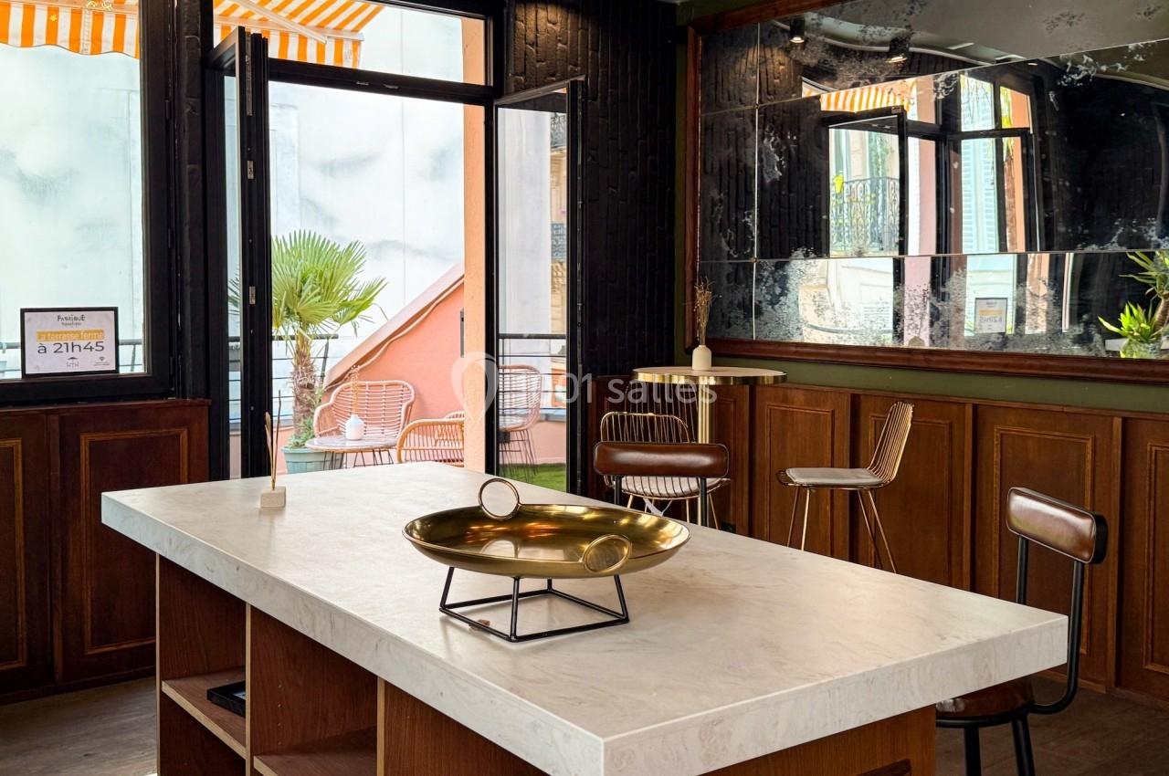 Intérieur lumineux avec un comptoir en marbre, tabourets en cuir, miroir mural et terrasse visible à travers une porte…