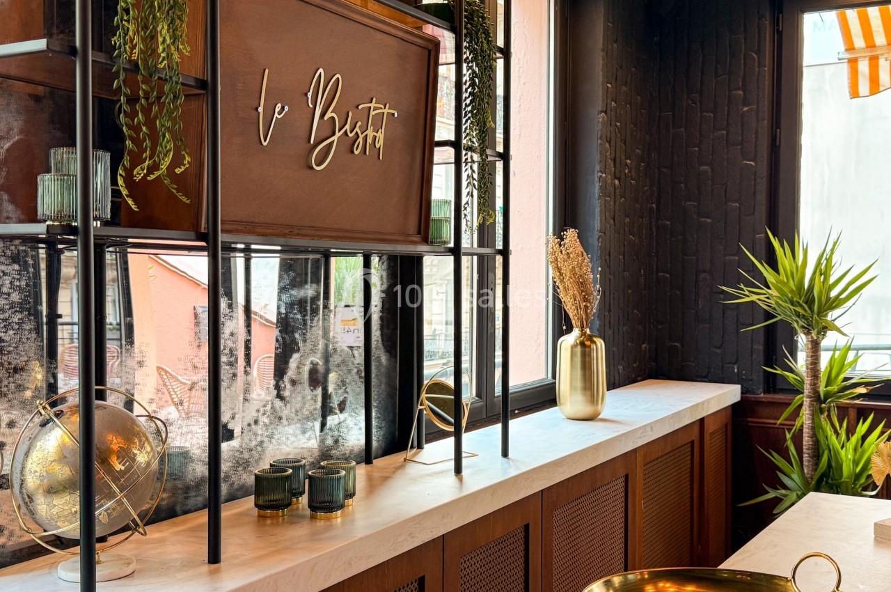 Étagère décorative avec plantes suspendues, globe terrestre, vases dorés et panneau ’Le Bistrot’ dans un intérieur lumineux.