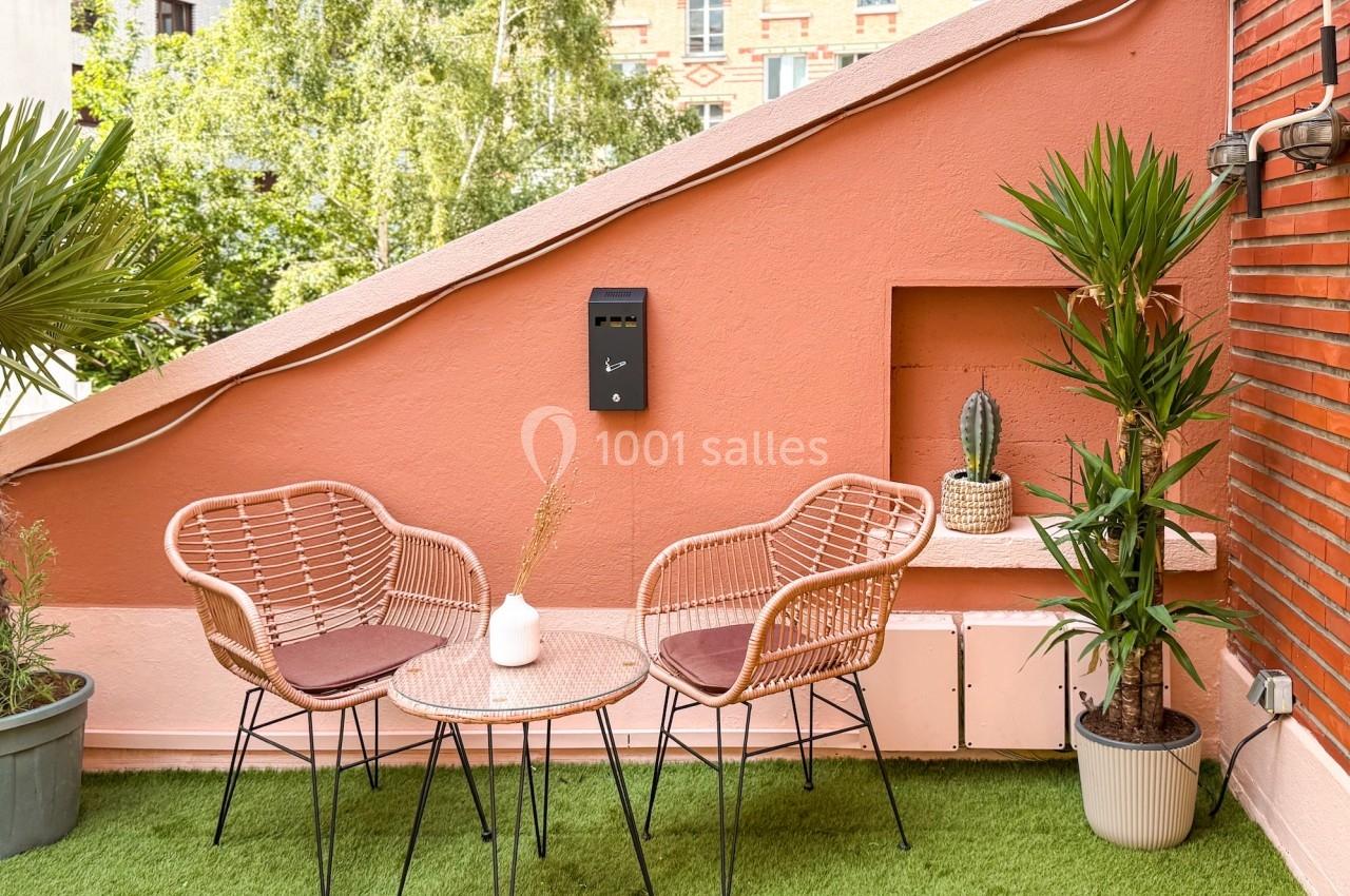 Terrasse aménagée avec table ronde, deux fauteuils en rotin, plantes en pot et mur rose en arrière-plan.
