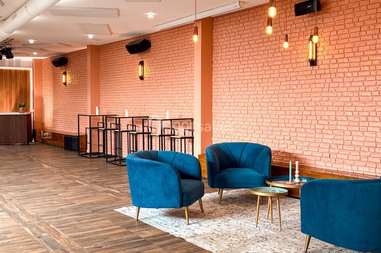 Espace intérieur lumineux avec murs en briques orangées, fauteuils bleus, tables basses dorées et éclairage suspendu.