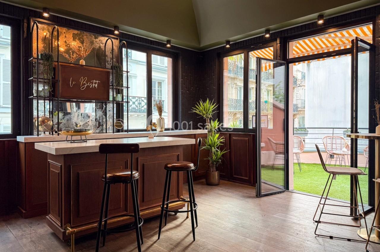 Intérieur d'un bar élégant avec comptoir en bois, tabourets, plantes et terrasse extérieure avec mobilier.