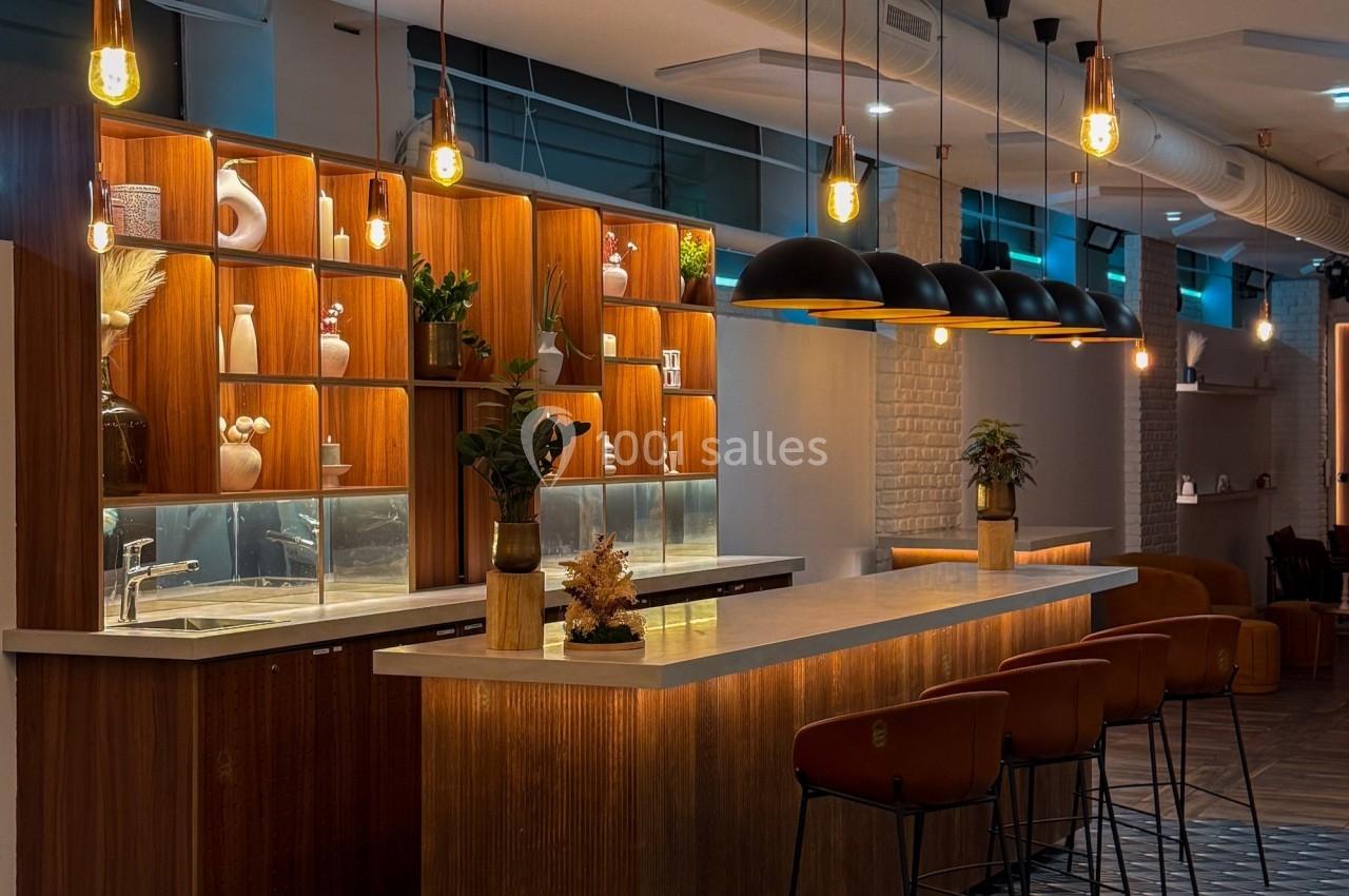 Bar moderne avec comptoir en bois, tabourets hauts, éclairage suspendu et étagères décorées d'objets et plantes.