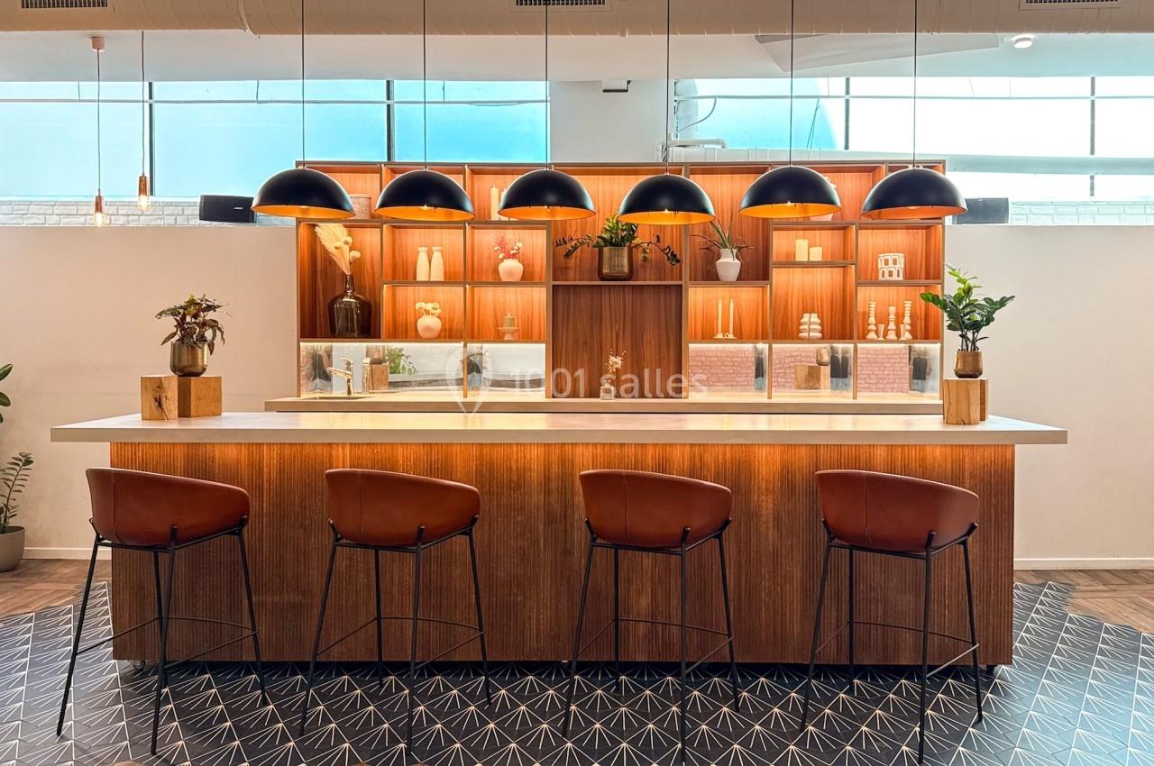 Comptoir en bois avec tabourets, étagères décoratives éclairées et suspensions noires dans un espace moderne et lumineux.