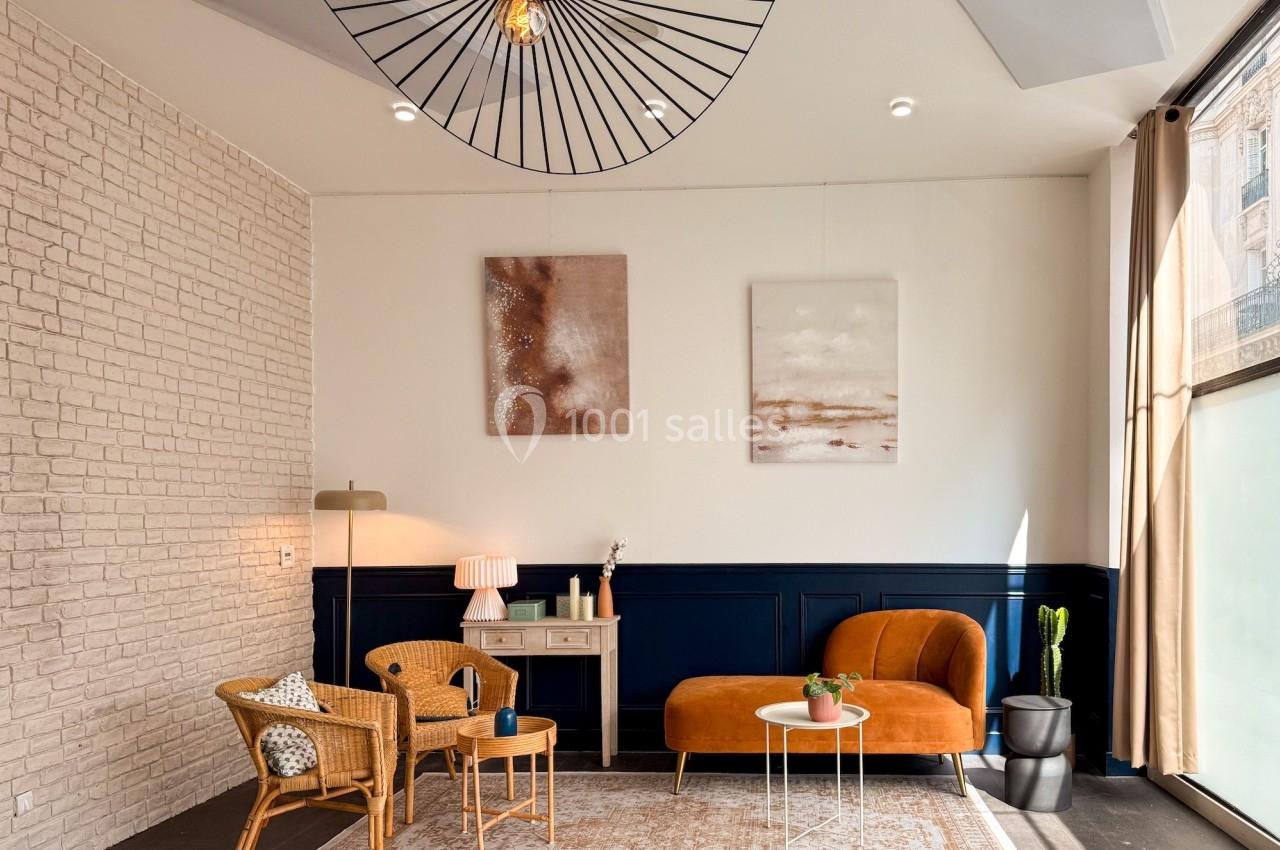 Salon lumineux avec murs en briques et bleu marine, mobilier en rotin et velours, tapis beige et grande suspension noire.