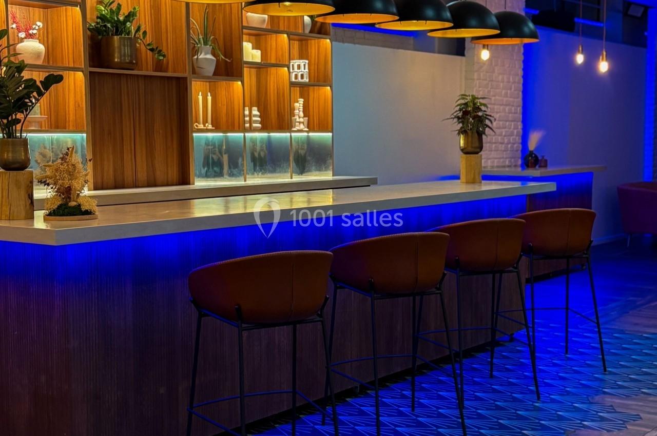 Bar moderne avec comptoir en bois, éclairage suspendu, tabourets en cuir et sol aux motifs géométriques bleus.