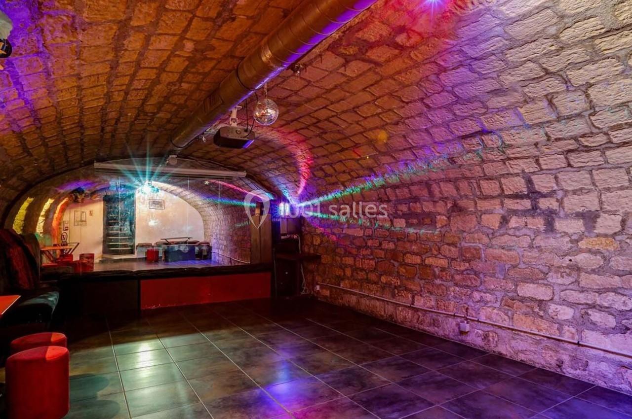 Salle voûtée en pierre avec éclairage coloré, petite scène et mobilier minimaliste.