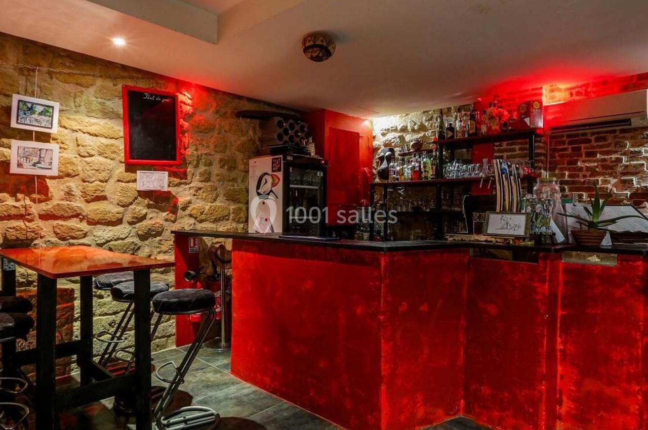 Bar avec comptoir rouge éclairé, tabourets hauts, murs en pierre et briques, décorations murales et étagères avec bouteilles.