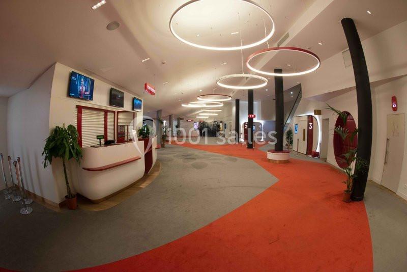 Hall d'accueil spacieux avec comptoir d'information, écrans, plantes décoratives et moquette rouge.