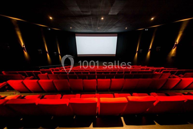 Salle de cinéma vide avec des rangées de sièges rouges et un grand écran blanc éclairé.
