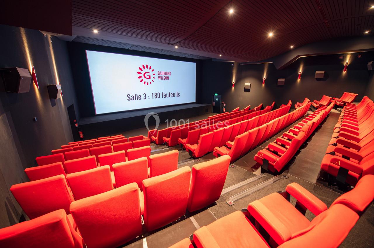 Salle de cinéma moderne avec sièges rouges vides, écran affichant ’Salle 3 : 180 fauteuils’ et éclairage tamisé.