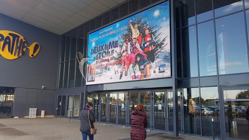 Affiche d'un film projetée sur un écran géant à l'entrée d'un cinéma Pathé, avec des passants devant.