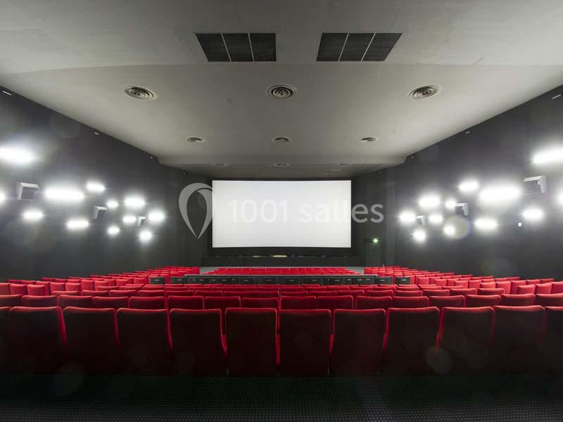 Salle de cinéma vide avec des rangées de sièges rouges et un grand écran blanc éclairé.