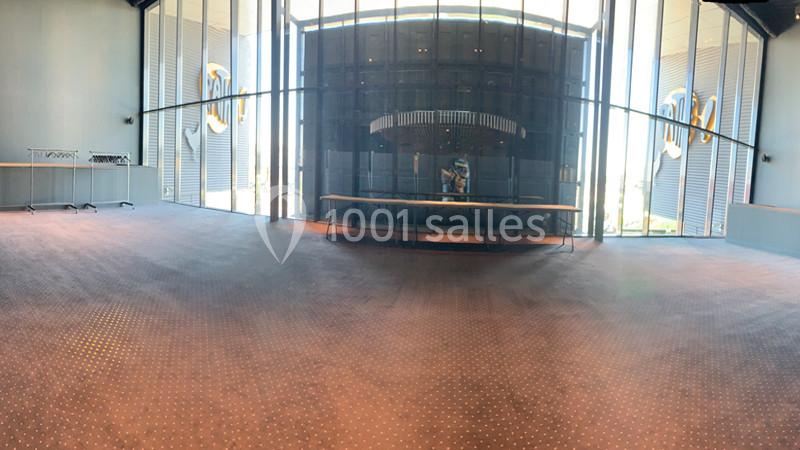 Salle vide avec moquette sombre, grande baie vitrée et structures métalliques visibles en arrière-plan.