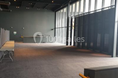 Salle vide avec moquette sombre, grande baie vitrée et structures métalliques visibles en arrière-plan.