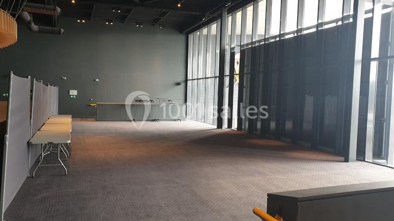 Salle vide avec moquette grise, cloisons amovibles, grandes baies vitrées et mobilier minimaliste.