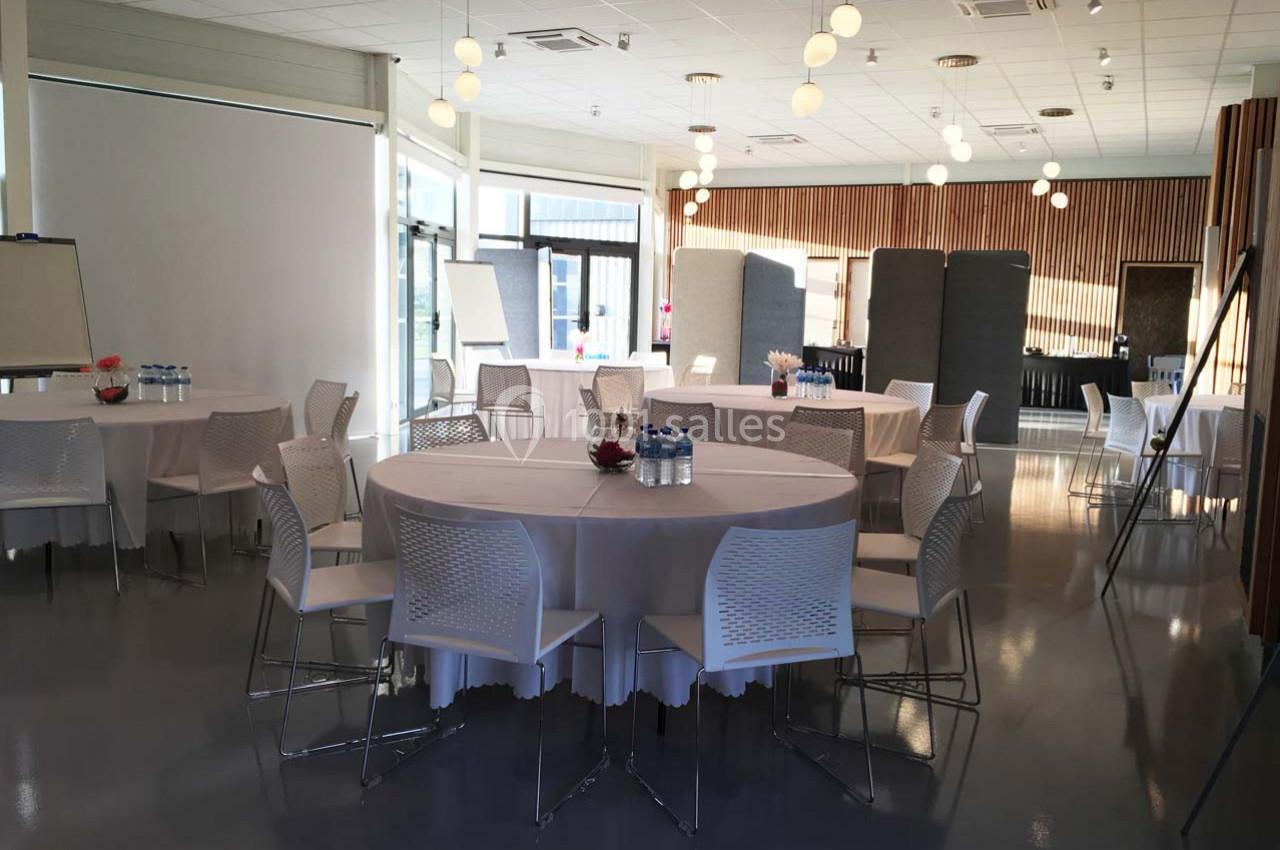 Salle lumineuse aménagée avec des tables rondes, des chaises blanches et des équipements pour une réunion ou un événement.