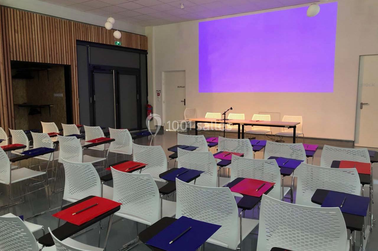 Salle de conférence avec des rangées de chaises blanches équipées de tablettes, face à un écran de projection violet.