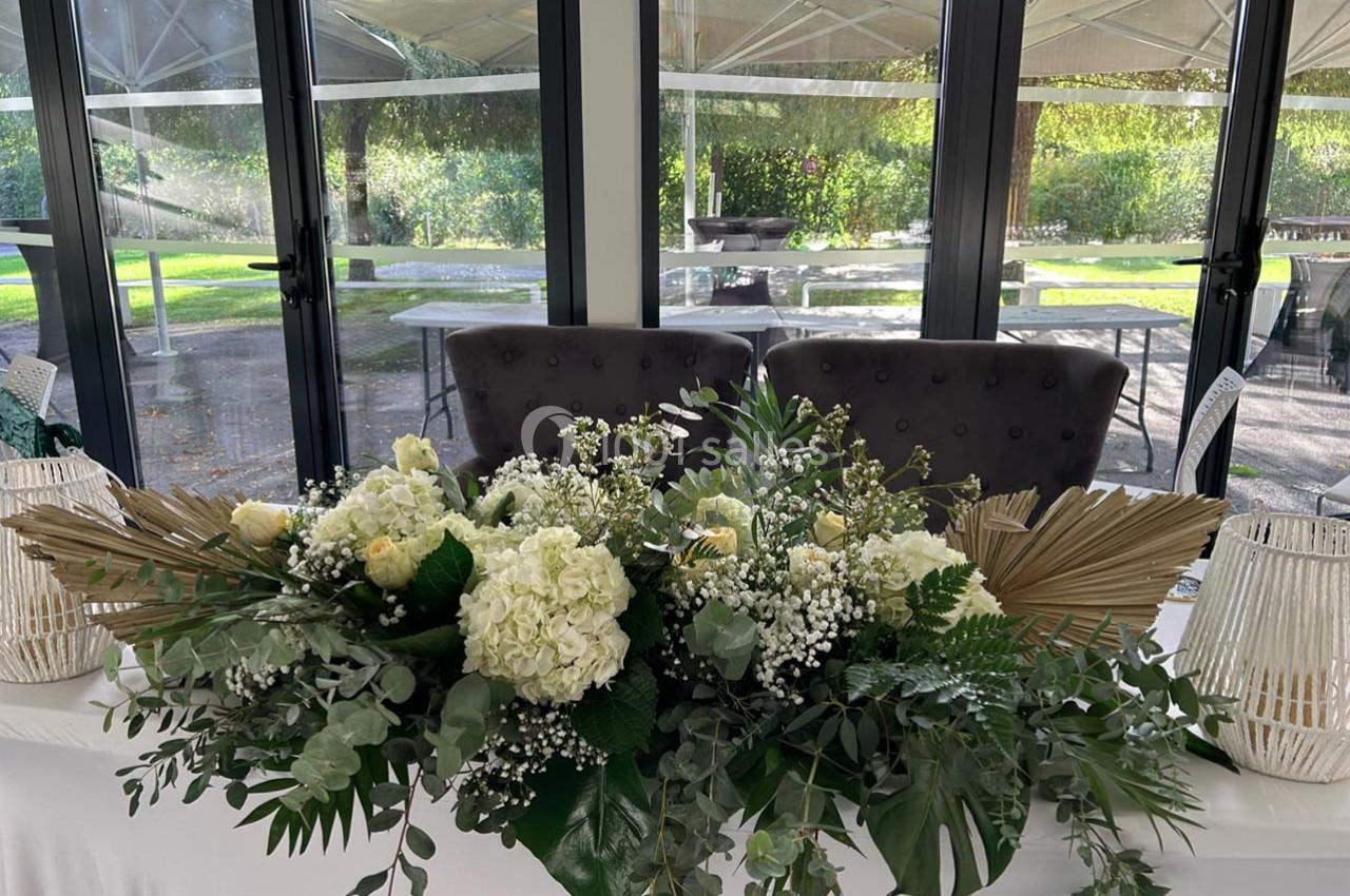 Composition florale blanche et verte avec des feuillages et des éléments décoratifs, disposée sur une table devant des baies…