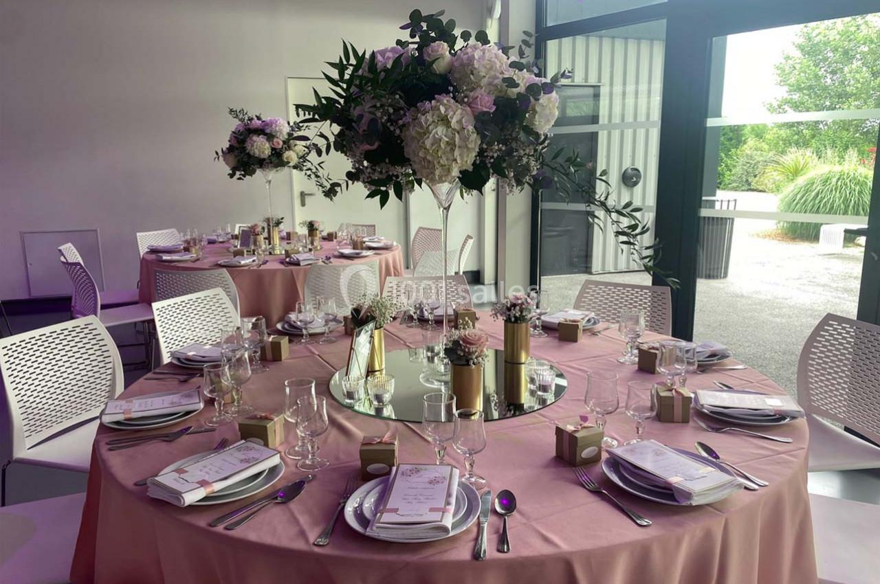 Salle de réception avec tables rondes décorées de nappes roses, centres de table fleuris et couverts élégants.