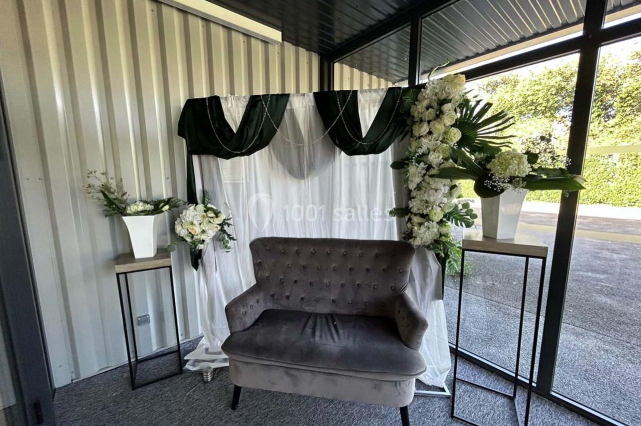 Canapé gris devant un rideau blanc décoré de drapés verts et de compositions florales blanches sur des supports.