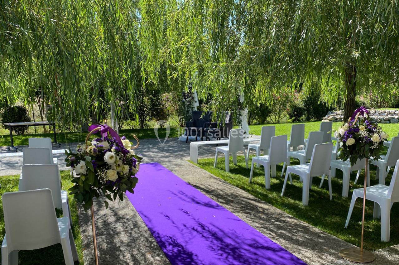 Allée centrale avec tapis violet, entourée de chaises blanches et décorée de fleurs, sous des arbres dans un jardin.