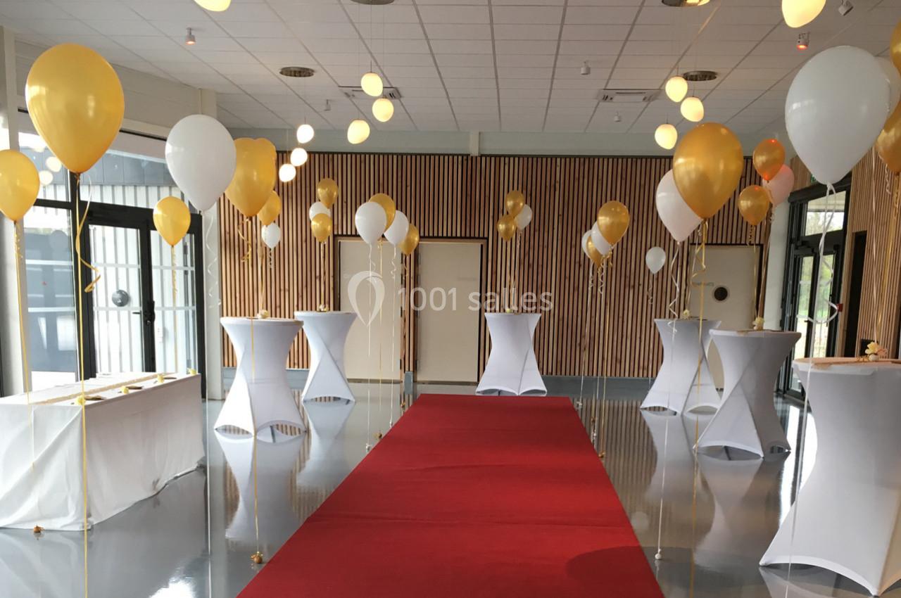 Salle décorée avec des ballons dorés et blancs, tables hautes drapées de blanc et tapis rouge au centre.