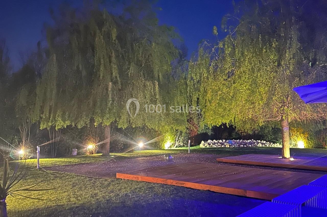 Jardin éclairé de nuit avec arbres, pelouse, terrasse en bois et bancs illuminés par des lumières tamisées.
