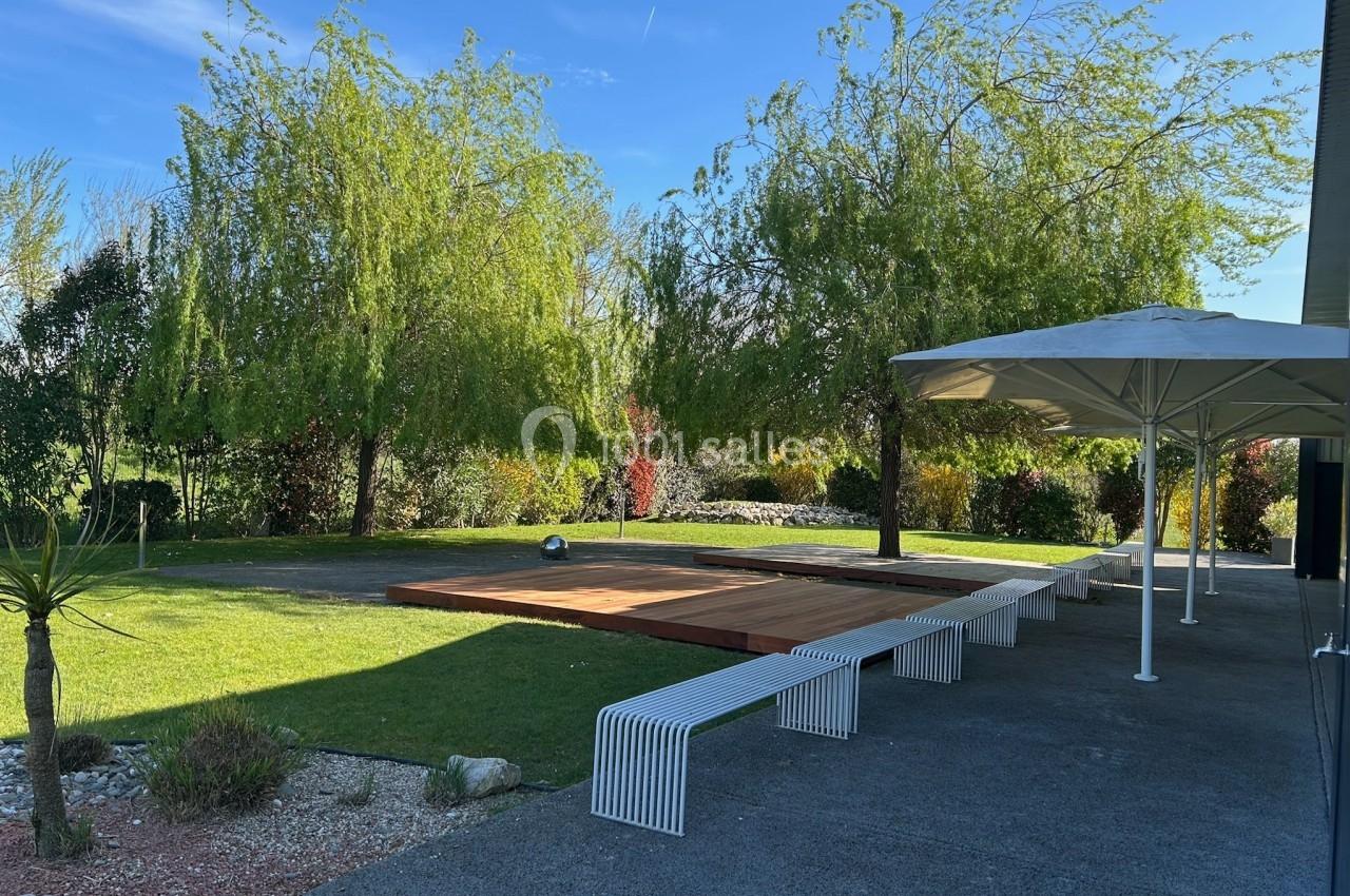 Terrasse en bois entourée de pelouse, arbres et bancs blancs, avec un parasol et un ciel dégagé en arrière-plan.