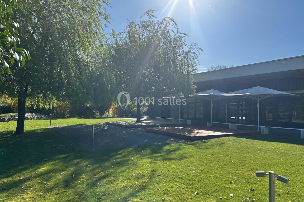 Pelouse ensoleillée avec arbres, terrasse en bois et parasols près d'un bâtiment moderne.