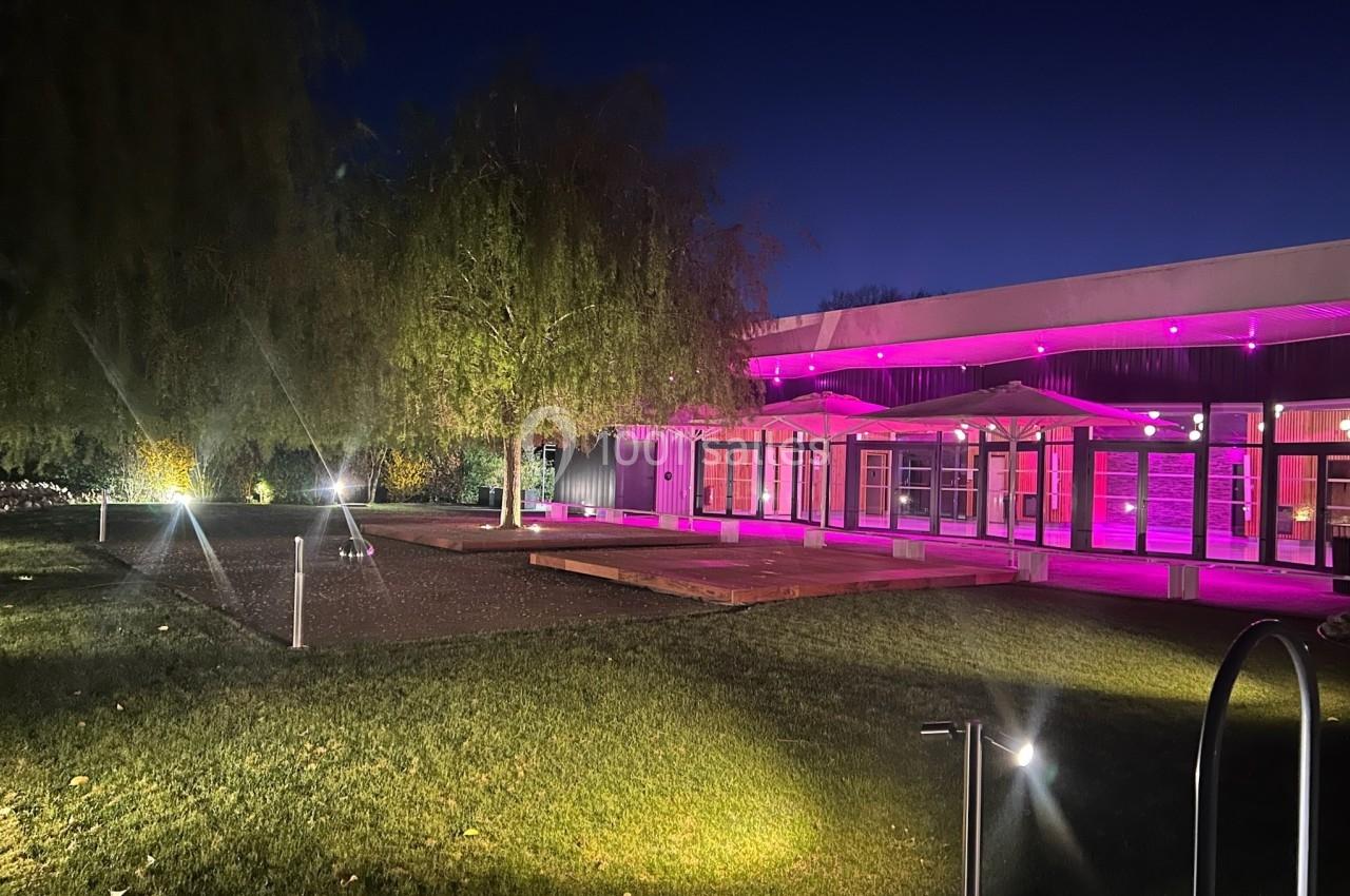 Bâtiment moderne éclairé par des lumières roses, entouré d'un jardin avec des arbres et un éclairage au sol, de nuit.