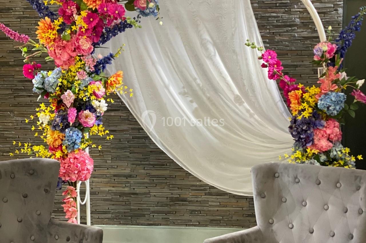 Arche décorative ornée de fleurs colorées et drapée blanche, encadrée par deux fauteuils gris devant un mur texturé.