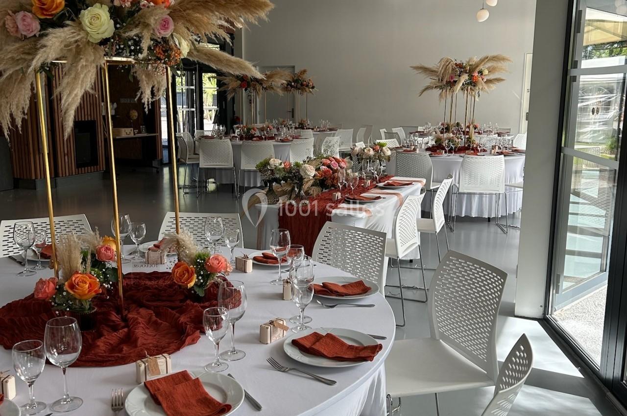 Salle de réception décorée avec des tables rondes, nappes blanches, serviettes orange et centres de table fleuris.