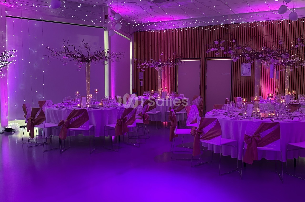 Salle de réception dressée pour banquet avec tables rondes mauve clair, chaises noires, éclairage violet et guirlandes lumineuses.