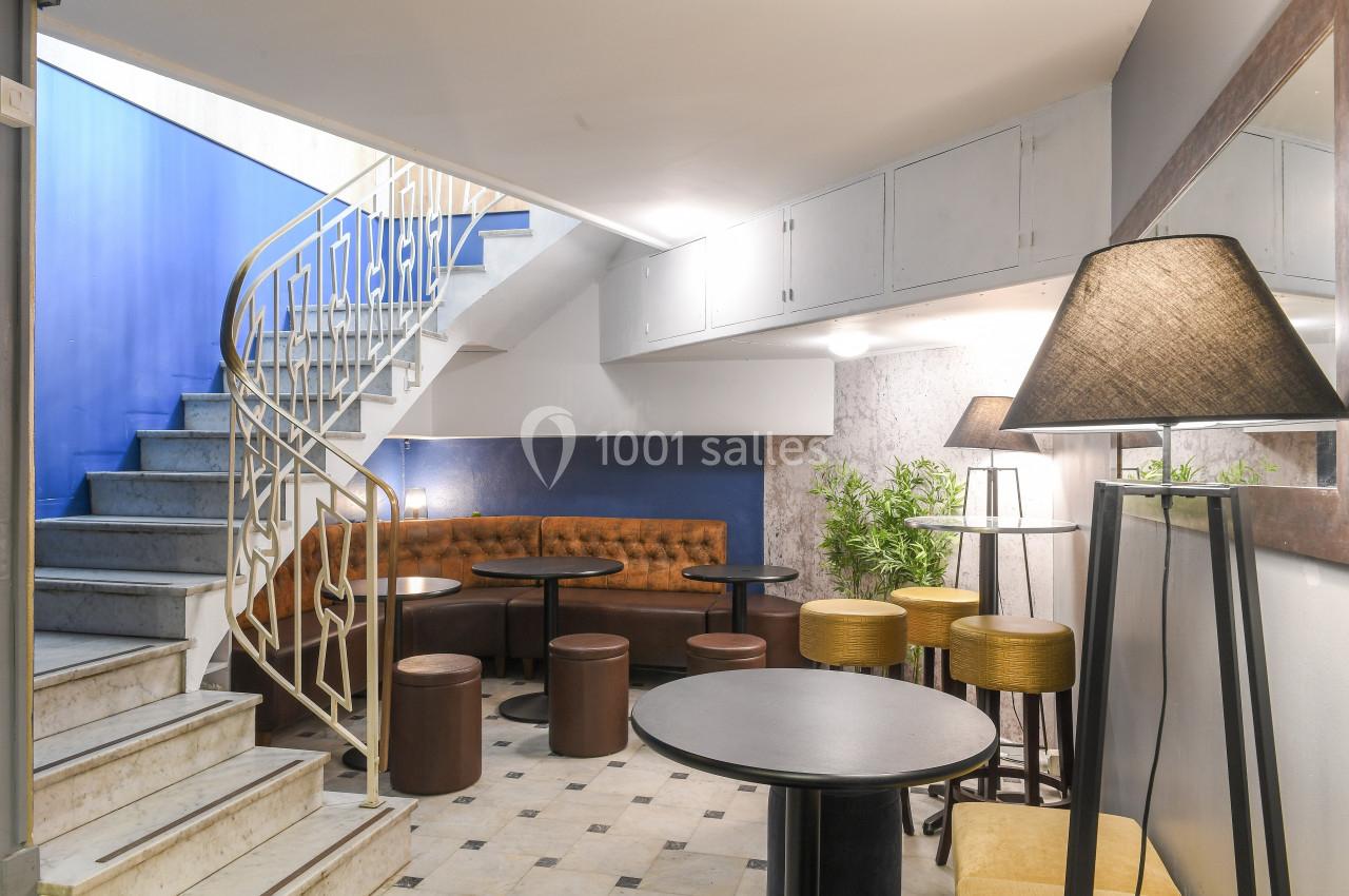 Espace intérieur avec escalier, tables, tabourets, banquette en cuir et éclairage tamisé, ambiance moderne et conviviale.