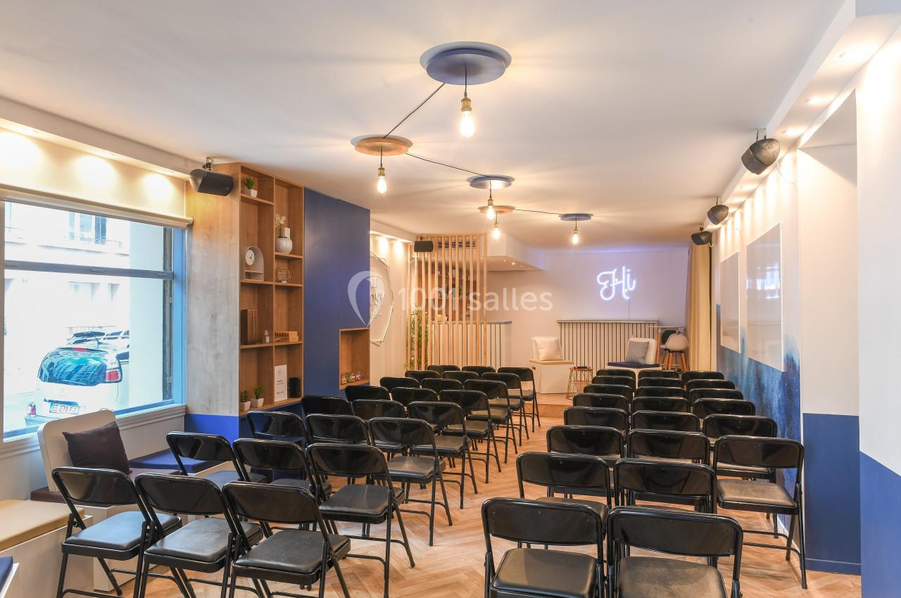 Salle lumineuse avec des rangées de chaises noires, un éclairage suspendu et un mur décoré d'un néon ’Hi’.