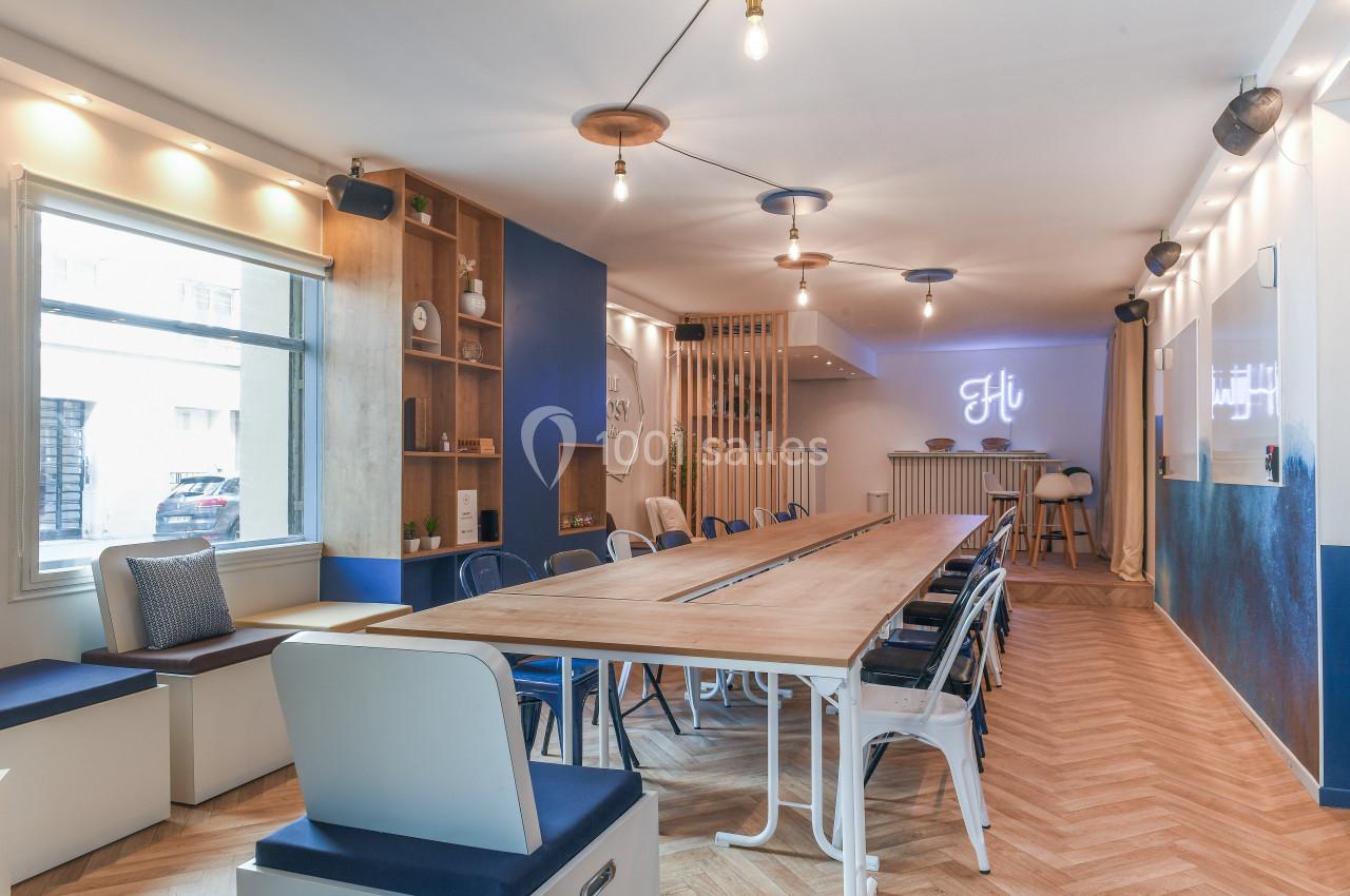Salle lumineuse avec grande table en bois, chaises variées, étagères décoratives et éclairage suspendu moderne.