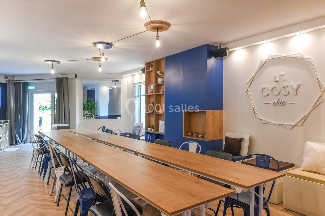 Salle lumineuse avec une grande table en bois, chaises variées, étagères décoratives et enseigne murale ’Le Cosy Chic’.