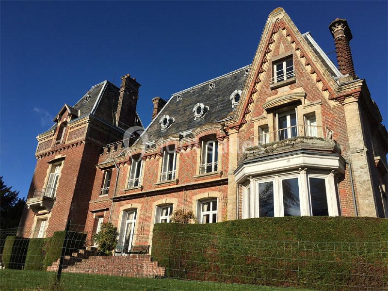 Grande maison en briques rouges avec toit en ardoise, entourée d'une haie et d'une clôture, sous un ciel bleu.