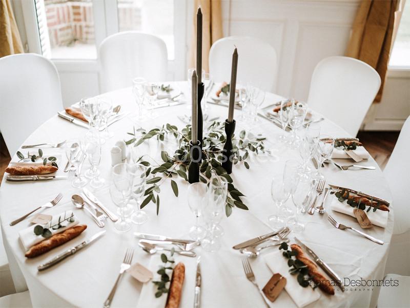 Table ronde élégamment dressée avec nappage blanc, bougies noires, feuillage central, couverts et baguettes individuelles.