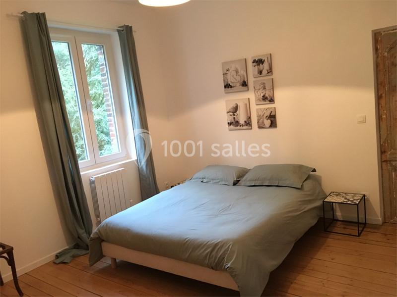 Chambre lumineuse avec un lit double, draps gris, rideaux verts, parquet en bois et décorations murales.