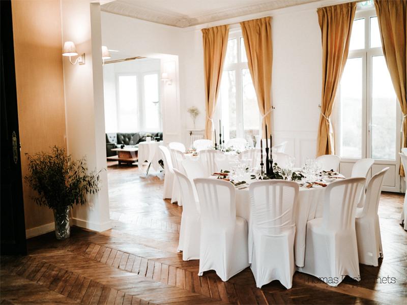 Salle de réception lumineuse avec tables rondes dressées, chaises blanches et rideaux dorés encadrant de grandes fenêtres.