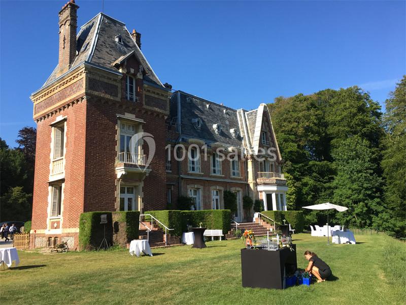 Manoir en briques rouges avec toit en ardoise, entouré de pelouse, tables blanches et arbres en arrière-plan.