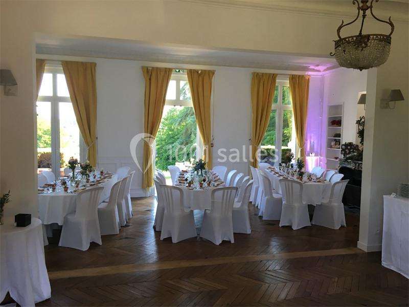 Salle lumineuse aménagée pour un repas, avec tables rondes décorées, chaises blanches et grandes fenêtres donnant sur un…
