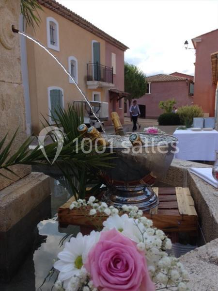 Seau en métal rempli de bouteilles de champagne dans une fontaine, décoré de fleurs et entouré de bâtiments colorés.