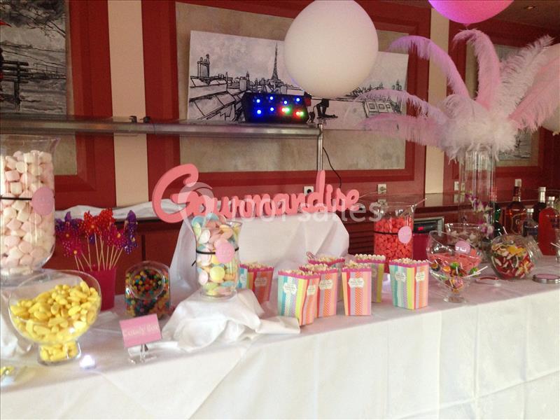 Table décorée avec des bonbons colorés, des pots en verre, des popcorns et une pancarte ’Gourmandise’ dans un cadre festif.