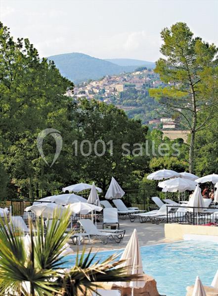 Piscine extérieure entourée de transats et parasols, avec vue sur un village perché et des collines boisées.